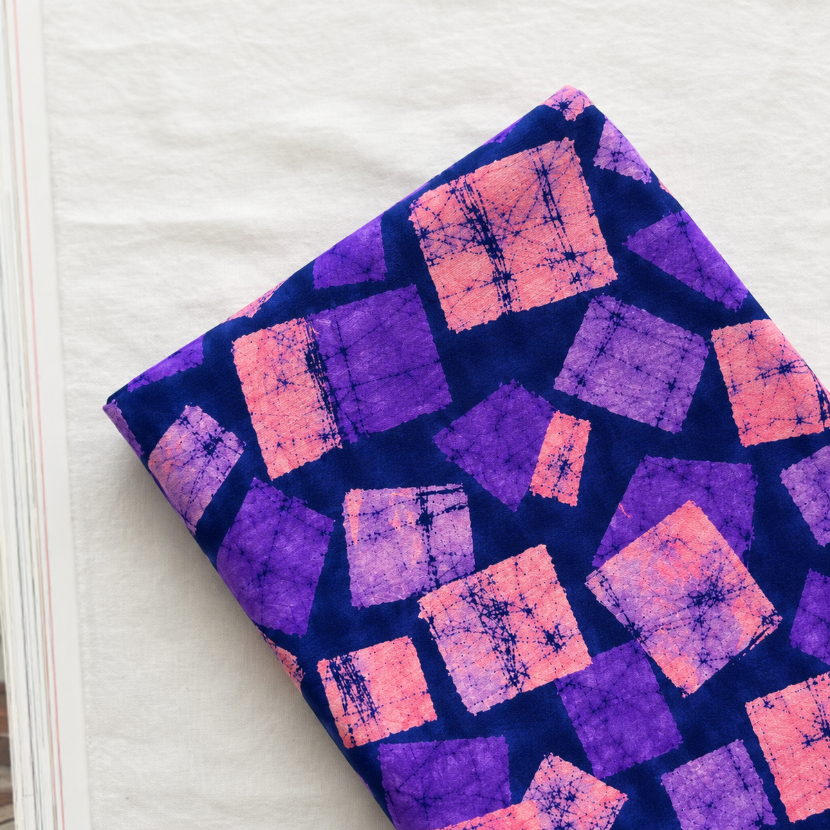 A folded piece of fabric with a vibrant purple and pink tie-dye geometric pattern, displayed diagonally on a white textured surface.